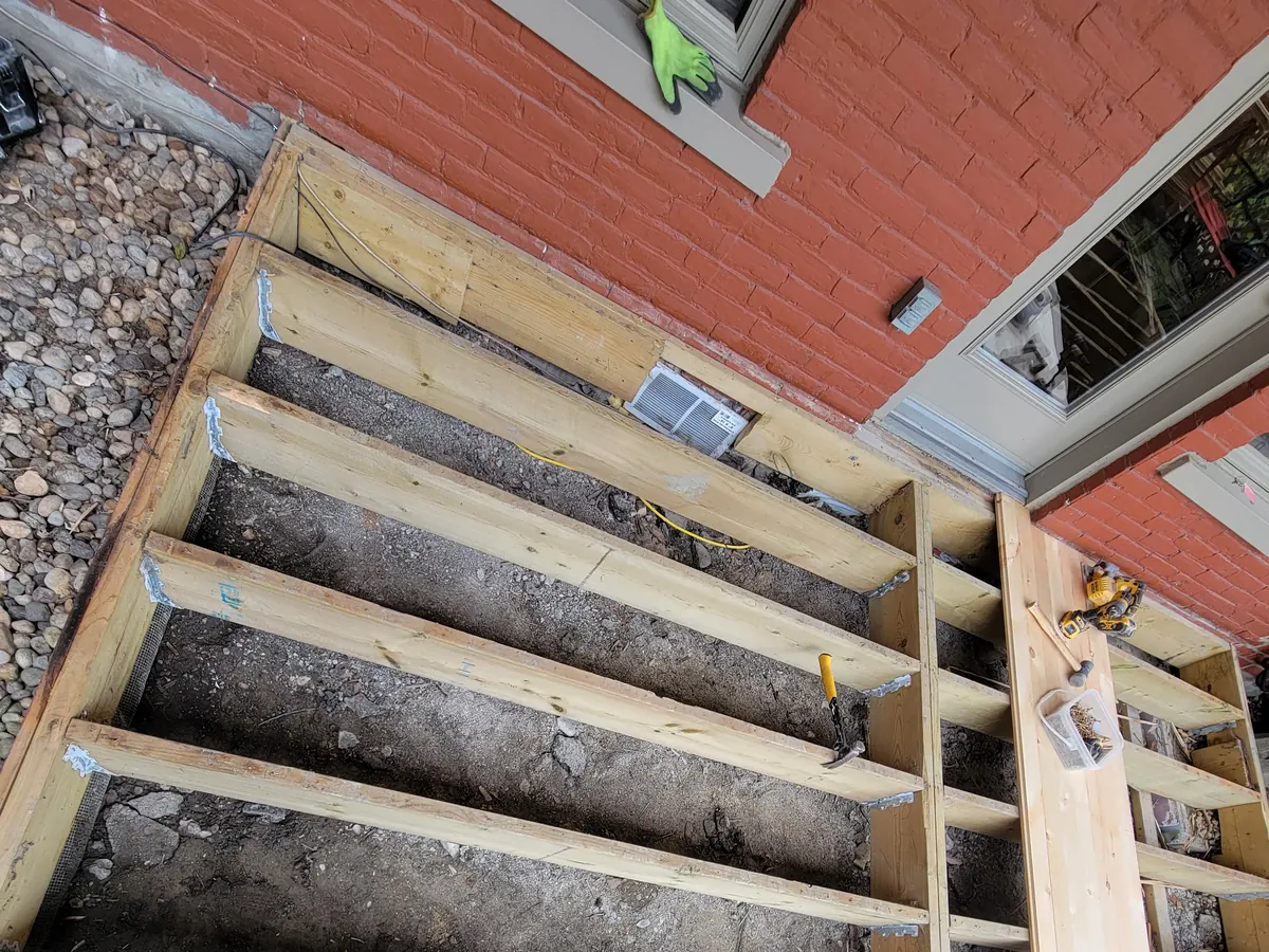 La structure de la terrasse en construction : solives neuves posées sur le sol de terre, contre le mur de brique de la maison